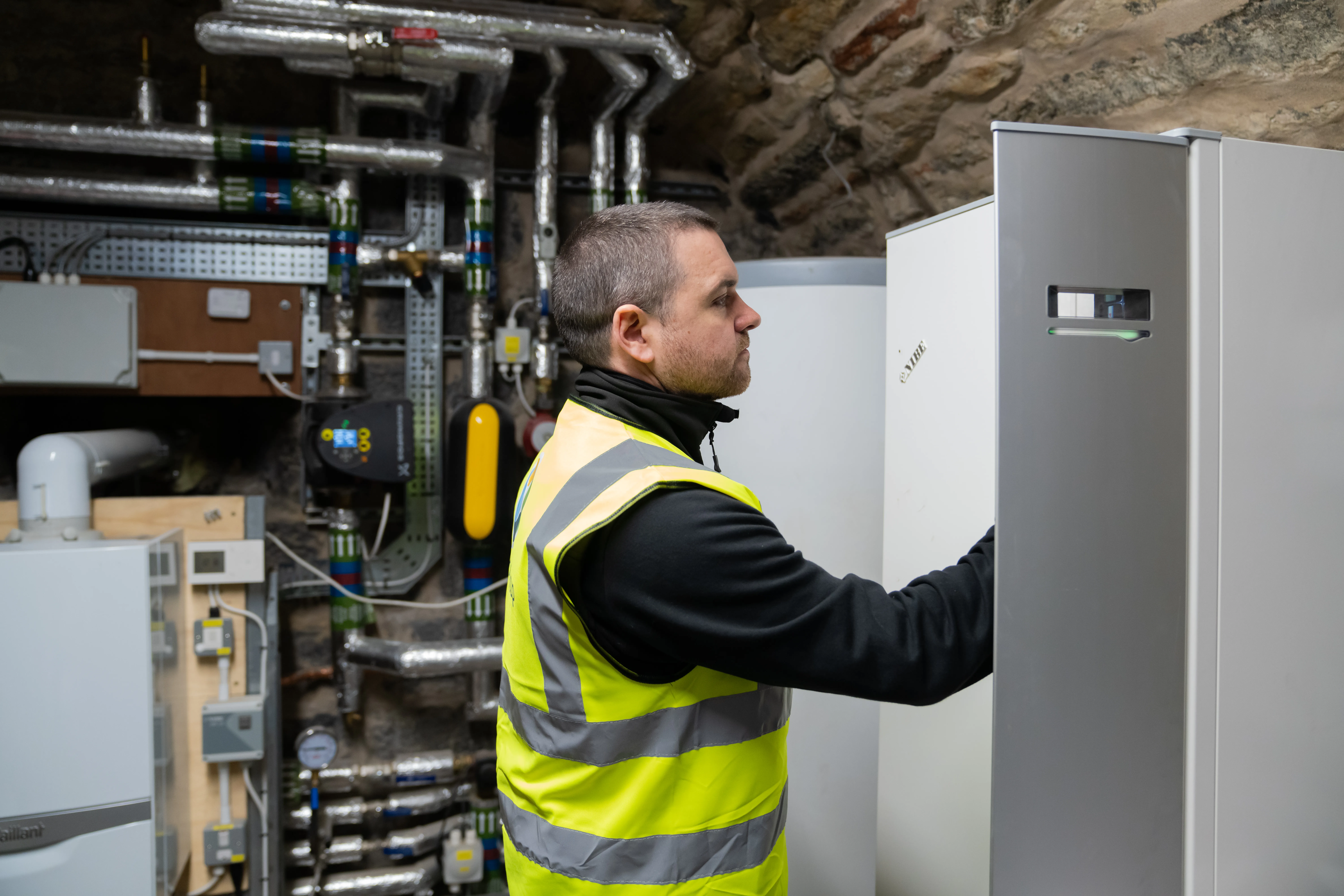McInnes Group engineer inspecting a completed air source heat pump installation — Vaillant boiler, NIBE heat pump cylinder, and detailed pipework against an exposed stone wall